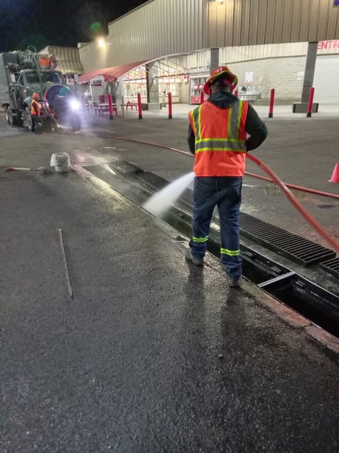 Worker in orange safety vest spraying water on parking lot at night near commercial building