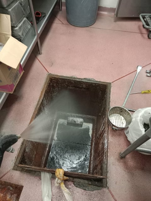 Open grease trap in commercial kitchen floor with steam rising, surrounded by cooking equipment and utensils