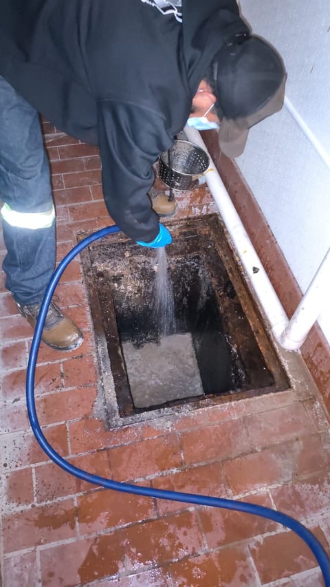 Person using a handheld strainer to drain water from a brick basement drain opening with a blue hose nearby