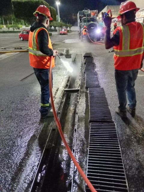 Two construction workers in safety gear cleaning a drainage channel at night with a water jet hose on a paved street