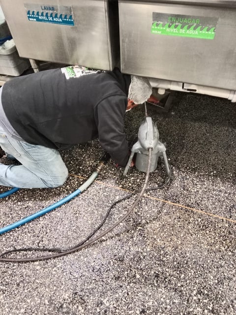 Worker in black clothing crouched down connecting a blue hose to water disposal containers labeled in Spanish